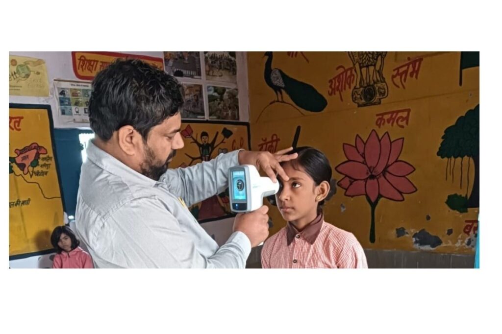 Photo Credits: Samyak
A man is using the eye-screening device to check the vision of a young school girl in a classroom setup with yellow walls